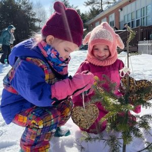 Two young students at Northern Lights School place birdseed ornaments on a young tree outside the school building.